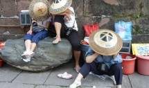 Title: Fenghuang Respite Caption: Three women huddled in the shade of their doulis for a brief rest along the banks of the Tuo River. With cameras around their necks, rental costumes stashed in plastic buckets, and laminated sample photographs propped against the ancient city wall, they took a break from tourist photography in the afternoon heat. Tourism has become an important part of the rapidly changing economy of Fenghuang County in Hunan Province, China. For the local Miao and Han peasant women, tourism has meant an opportunity to earn a small income by dressing tourists in mock ethnic costumes and taking their photographs. Yet this day business was slow as few tourists strolled the waterfront in the heat.