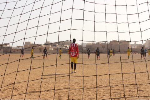 View of a game unfolding on a soccer pitch from behind the goal's net, with the goalkeeper in red standing on the other side. 