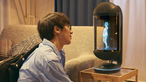 A young man in glasses sits on the floor in front of a small endtable that's beside a sofa. Entranced, he looks at a character projection of Azuma Hikari. She's about 10 inches tall, wearing a holter top white dress, with above the knee white and blue striped socks, and she strikes a flirtatious pose.