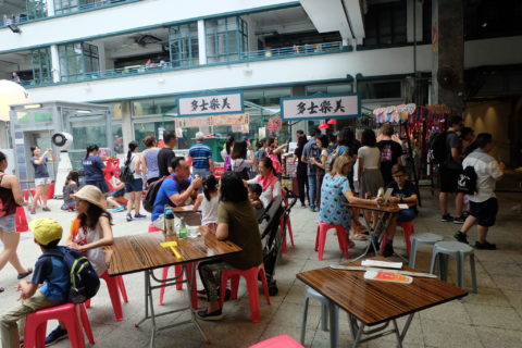 A crowd of people move through what seems to be the courtyard of a mall, with families sitting at tables in the foreground and throngs of people going in and out of stores in the background.