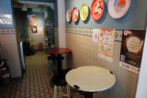 The inside of a restaurant, with tall, round tables, an accletic set of stools, and a row of plates, each painted with different flowers hanging on the wall.