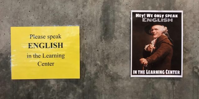 The photograph displays two signs. The sign on the left has a yellow background and black letters that read "Please speak ENGLISH (in all caps) in the Learning Center." The sign on the right is a photograph of a man, possibly of Anglo-Saxon descent, who is wearing Renaissance-style clothing and smiling. He is pointing at the viewer and the accompanying text reads, in all caps, "HEY! WE ONLY SPEAK ENGLISH IN THE LEARNING CENTER."