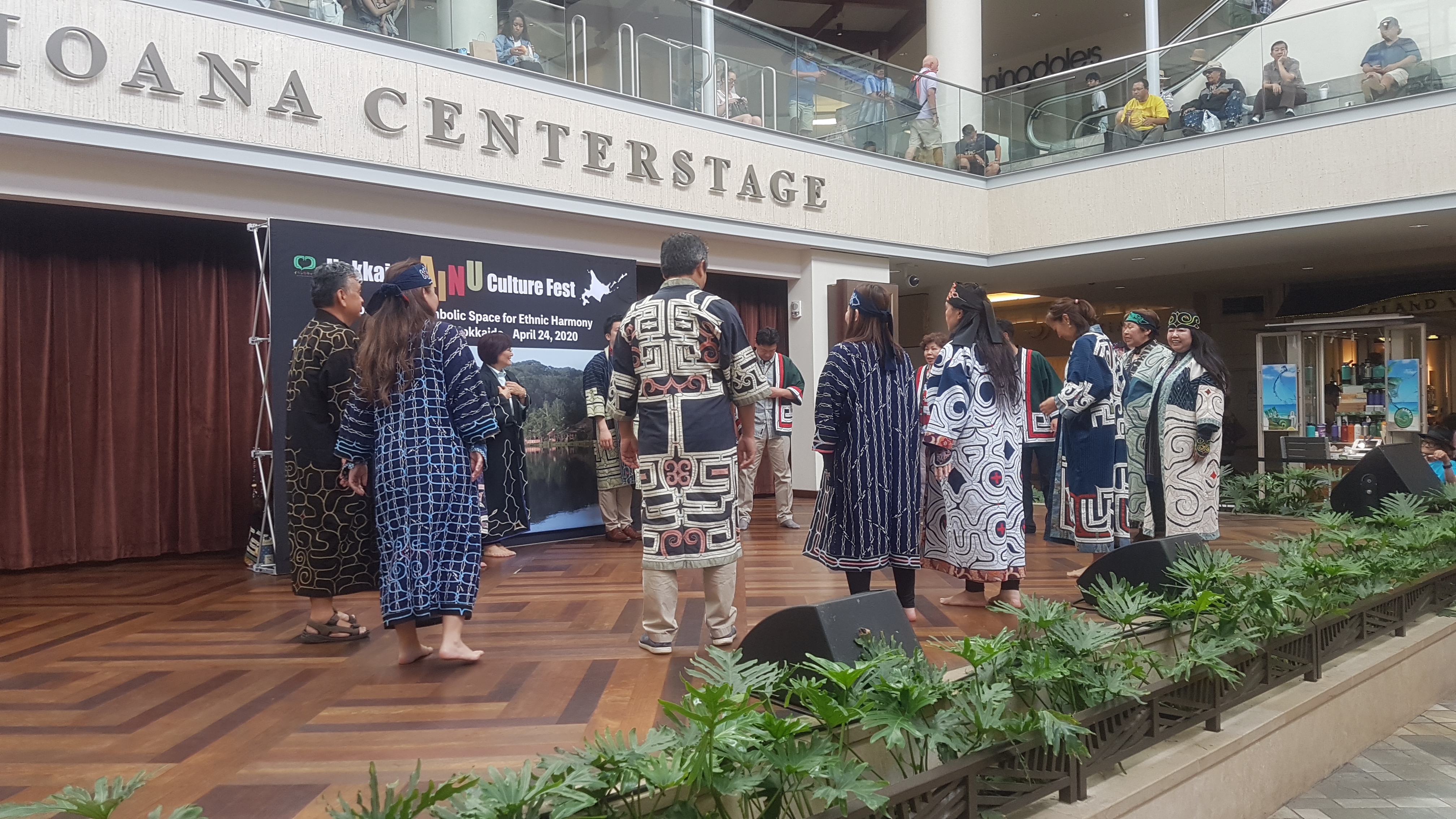 About thirteen men and women dressed in traditional embroidered dark blue robes form a circle on a wooden stage with the words "Ala Moana Centerstage" on the second floor overhang. The words are cut off and only "centerstage" can be seen. Some audience members are looking at the performance from the second floor of the mall.