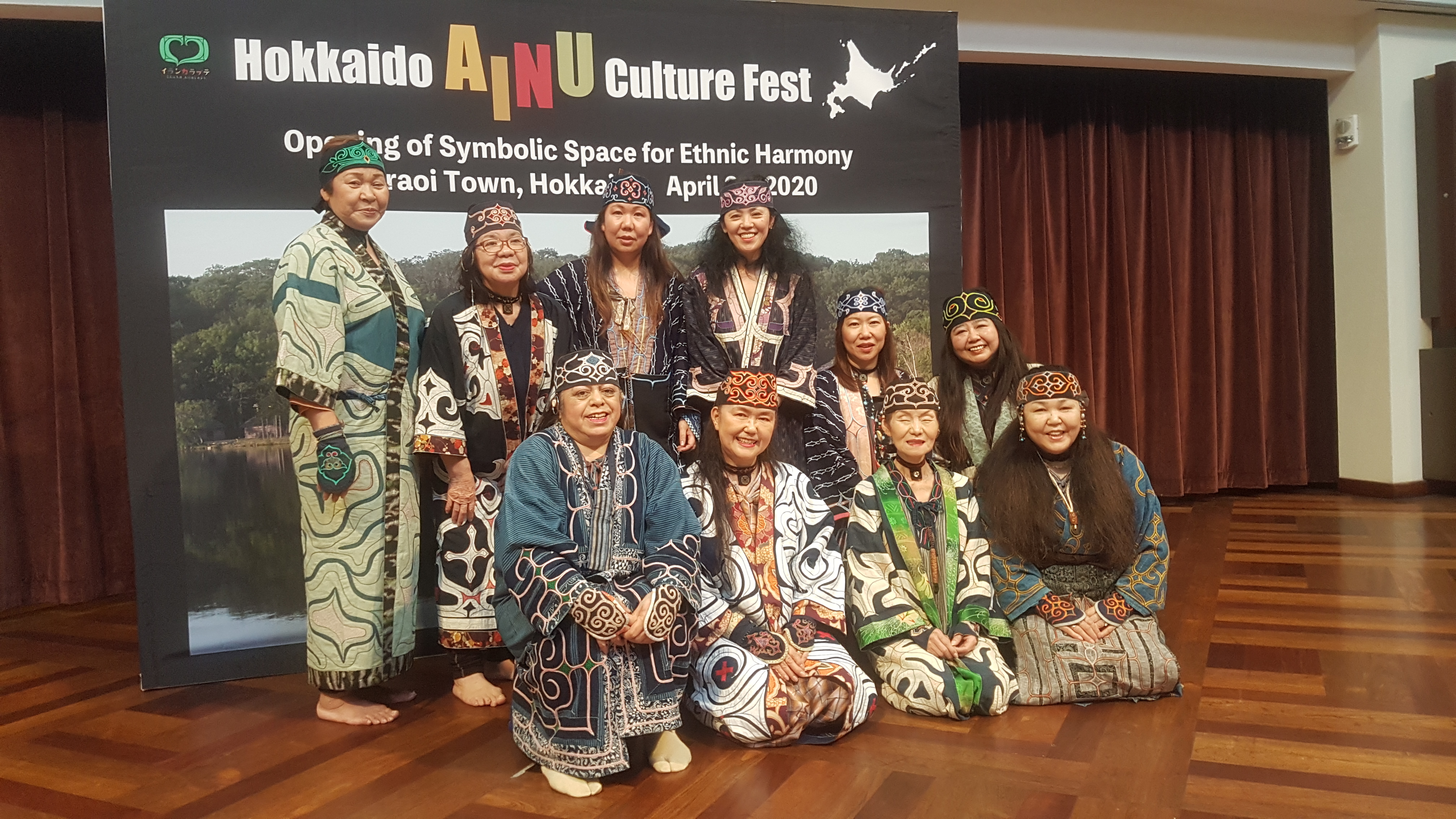 The ten women from the performance group poses on the stage, in front of a poster with the words "Hokkaido Ainu Culture Fest." Words in smaller fonts, with parts blocked by the standing performers, read "Opening of Symbolic Space for Ethnic Harmony, Shiraoi Town, Hokkaido, April 24, 2020." The back row has four women standing and two kneeling on the right, and the front row has four women kneeling. They are dressed in dark blue robes embroidered in colorful threads and cloths.