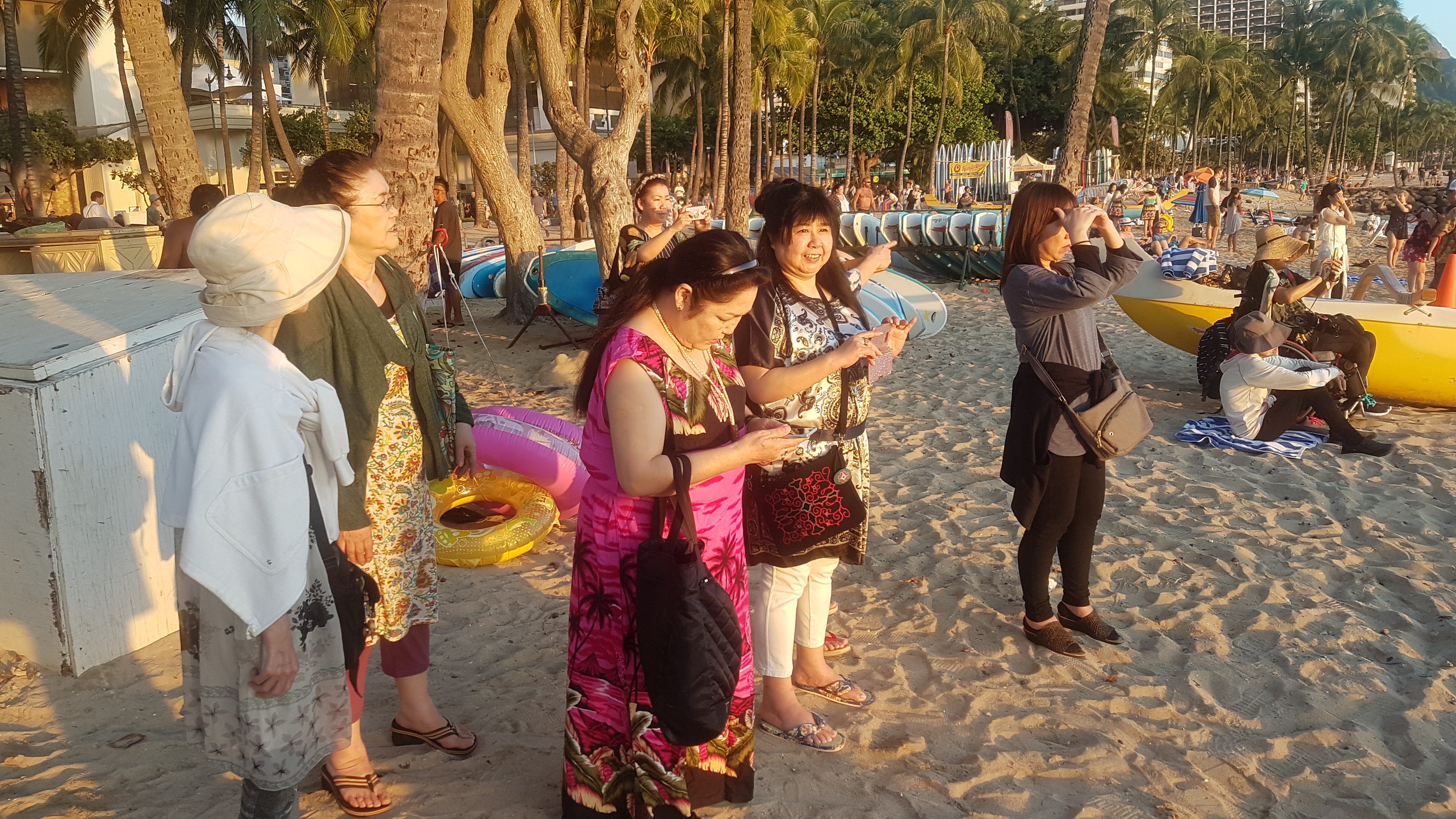 Six women dressed in summer wear on a beach with trees, surfboards and other tourists in the background. Some are gazing at the sea while some are in various stages of taking photographs with their mobile phones. The setting sun lights up their faces.