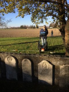 A young person moves over grass near farmland with a radar device, all behind four unmarked gravestones.