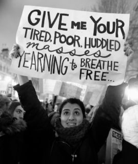 A woman holds a sign at a protest. The sign reads "Give me your tired, poor, huddled masses yearning to breathe free..."