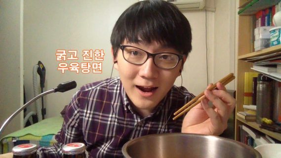 A young man holding chopsticks leans over a large phone bowl on the right, with a microphone jutting into the frame on the left.
