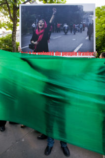 The sihlouette of a demonstrator behind a plain green banner holds a sign up over it.