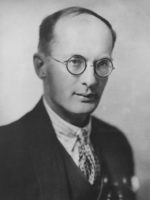 Photo portrait of man in suit and tie. He is balding and is wearing round wire spectacles.