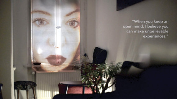 A woman's face is projected onto the window shade of a small darkened apartment. The text in the photo, a quote from an interlocutor, reads: "When you keep an open mine, I believe you can make unbelievable experiences."