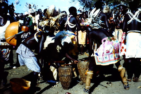 Men in the foreground enthusiastically lean over to play drums. On the other side of them a circle of men in ritual garb dance.