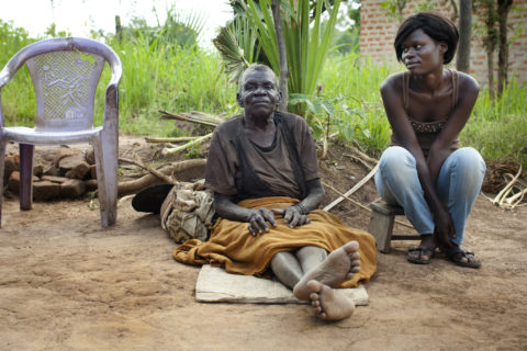 A Grandmother sits on a mat outside, her legs extended. On her left side, her grown grandaughter sits on a short stool.