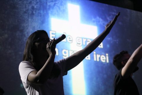 A woman speaking into a microphone and holding one hand in the air, palm up. A screen behind her has an illuminated cross and words in Portuguese.