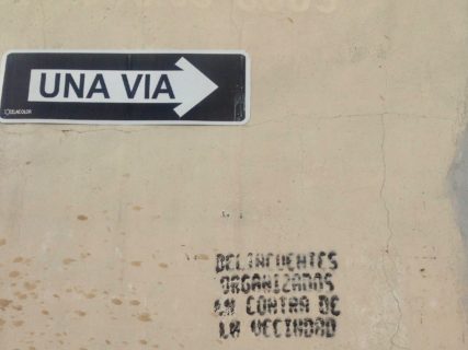Cement wall with a street sign reading "una via" (one way) affixed to it. The stencil is written below.