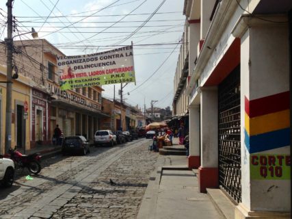 A city street with the silhouette of one pedestrial in the distance. The treet is lined with parked cars and motorcycles.