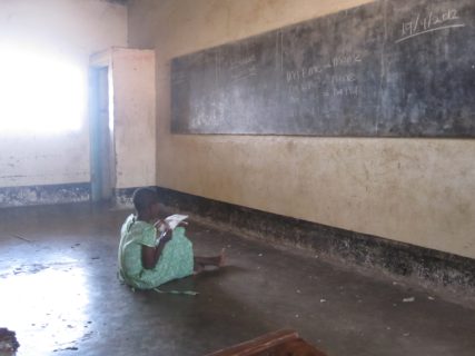 A female student sits on the floor of a classroom, looking up at a chalkboard and taking notes.