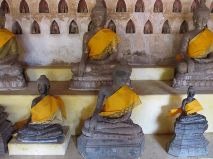 Photo shows six Buddha statues, three on a raised platform and three below. A scarf is draped over the left shoulder and wrapped under the right arm of each statue.