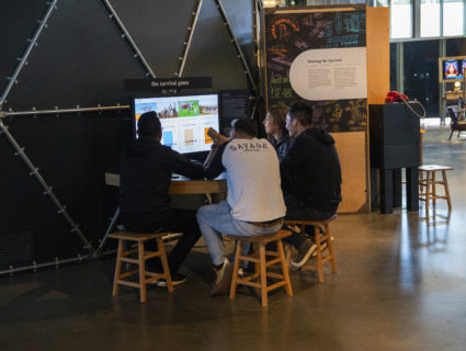 Three young people sit in front of the survival game console. One's hand is outstretched, pointing at the screen.