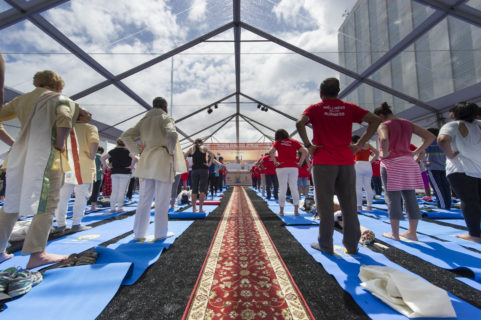 What looks like nearly 50 people stand barefoot on their yoga mats, lined up in a grid. The Ceilingis glass, showing a blue sky with large clouds. The room is drenchedin sinlight. Each person stands facing forward with their hands on their hips.