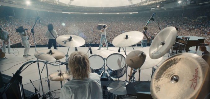 Scene of Queen's Live Aid concert that looks out from behind the drummer to show Freddy Mercury singing before a sold out stadiem.