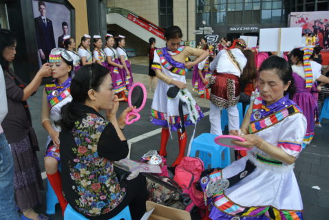 Three women are sitting on plastic stools: two put on their makeup and hold mirrors up to their faces. The third woman closes her eyes as a woman standing adjusts the headress of her costume. A fourth woman, who is standing, appears to be zipping up the side of her dress.