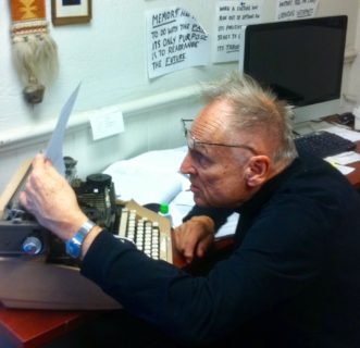 Roy Wagner sits in front of a typewriter, glasses perched on his forehead, leaning into the piece of paper that he's holding up to read.