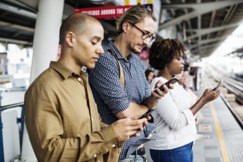 Three people standing near each other on a train platform, all staring intently at their smartphones. 