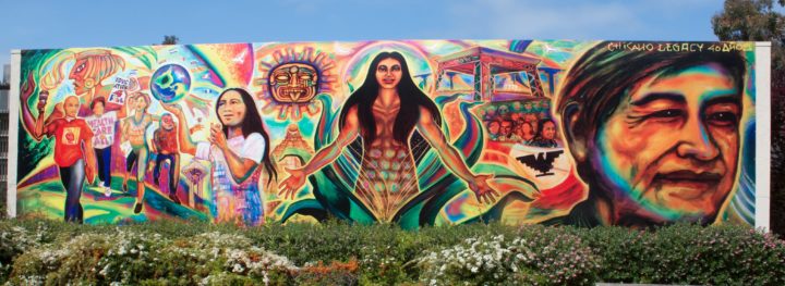 Large mural. In the center a woman emerges from the bud of a flower, her arms outstretched and welcoming the iblookers. Ancient Mayan and Aztec pyramids and suns apear in the background. In the fore ground are Latinx youth marching and playing.