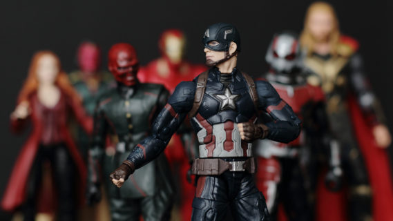 An action figure of Caption American stands front and center, posed with his fists clenched and ready to fight. Behind him and slightly out of focus, stand other action figures from the Marvel Universe