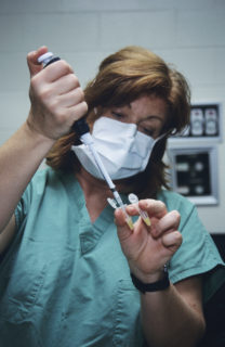 A woman in scrubs and wearing a surgical mask, injects fluid into a small tube held by her thumb and index finger. She has another tube clutched between her index finger and middle finger.