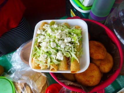 Corn tortillas that are rolled up (flautas) sit on a styrfoam plate, that is itself perched on a bowl of rolls. Lettuce, guacamole, and cheese are piled on top of the Flautas/ 