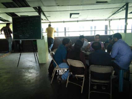 A group of people sits in folding chairs around a table. A callkboard full of writing stands to their right.