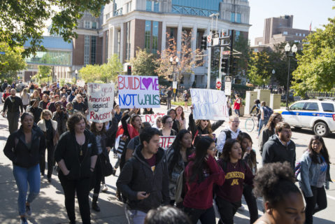 Fibonacci Blue, licensed under a Creative Commons Attribution License. A photograph of protesters of a range of races and genders marching with signs at the University of Minnesota. Some protesters are chanting. Some also wear University of Minnesota clothing. One sign reads "1st gen American against racism" and two others read "build love not walls."