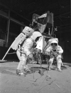Two men in space suits are bent over trying to solve a mechanical puzzle.