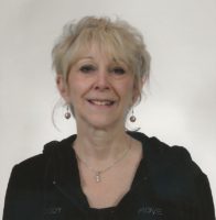 A white woman with blond hair that's pulled back smiles at the camera. She has bangs and dangly earrings. She wears a black hoodie sweatshirt.