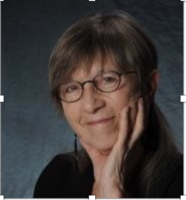 Portrait of a white woman with light-colored, whispy short hair. She wears glasses, and has ber hand on her chin.