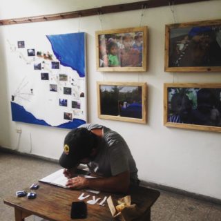 A man is hunched over a desk at the front of a room, cutting letters for what are likely stencils. The letters T-R-A are beside the paper the man is hunched over. On the wall behind him is a map of southern Mexico and Central America showing photos and a hand-drawn path.
