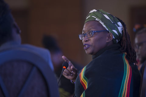 An African woman with glasses turns over her shoulder to a microphone , her right index pointing as she speaks. Her head is wrapped in a floral print, and her dredlocks are visible in the back.