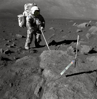 An astraunaut in a space suit is alone, standing on the surface of the moon. He is using a long contraption to sample the moon surface.
