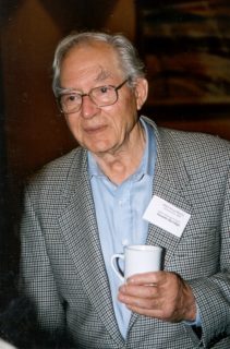 A pale man with classes and receding white hair wears a checkered suitjacket holds a cup of coffee. He has a nametag pinned to his collar.