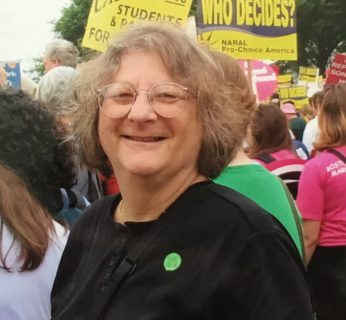 A white woman with glasses smiles at the camera. She wears a black t shirt and a round green pin. 