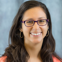 Portrait of a woman with dark hair that falls to her shoulders. She wears glasses and medium-sized hoop earrings.