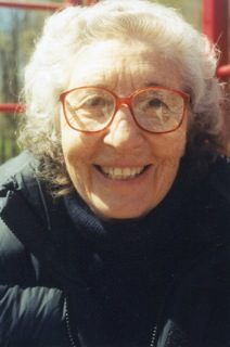 A white woman is smiling directly into the camera. She wears large-lensed glasses and she has white curly hair.