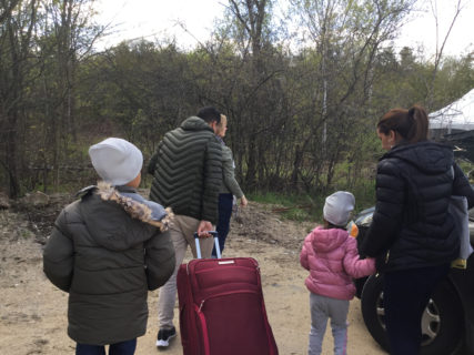 A family of four gets out of a car. They are all wearing large, puffy winter jackets. The father pulls a large suitcase behind him. The younger child clings to her mother. None of their faces are visible.