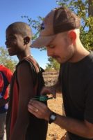 A young white man witha mustache is holding an unidentifiable object with his left hand and touching it with his right hand. He wears a weathered baseball hat. To his right, stands a young black man who is laughing.