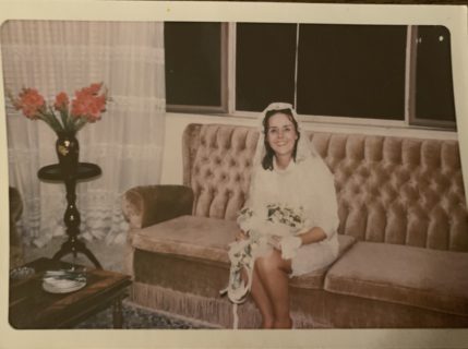 A woman with pale skin and dark brown hair sits on velvet a sofa. There are lard flowers of the sidetable to the left, and a window showing the night sky behind the woman. she appears to be in a knee-length wedding dress.