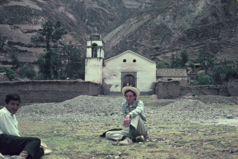 A small chapel sits at the base of a mountain. In front of it two men, one a white foreigner and the other a dark-skinned man, sit crossed legged on the ground.