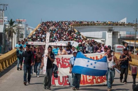 Thousands of people walk toward the camera over a white highway bridge. The people in front carry the national flag of Honduras, a cross that’s taller than the person carrying it, and a banner whose words are obscured by the men carrying the flag.