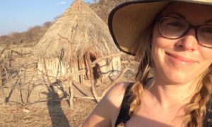 I white woman smiles at the camera. He face is shaded by a straw hat that tshe's wearing. She has glasses, and blond hair in two braids that fall past her shoulders. In the background is a straw hut surrounded by a wooden fence.